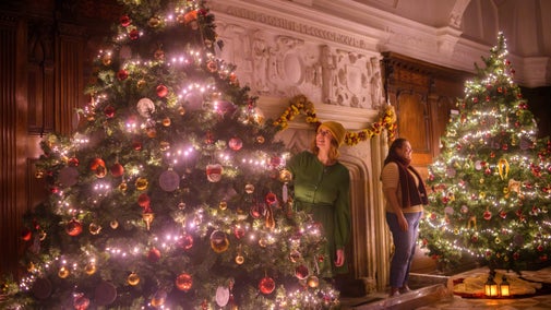 Two large Christmas trees decorated with red and gold baubles and fairy lights stand either side of a large fireplace. Two women are dressed in winter clothes admiring the trees. There is wood panelling and plasterwork in the background and lanterns on the ground.